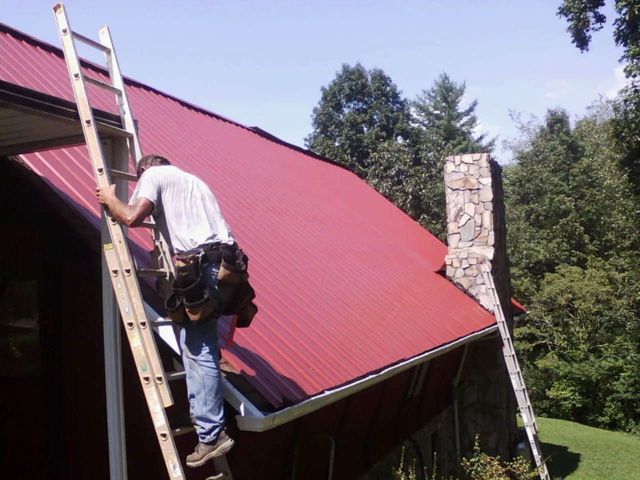Roofer on a ladder beside a red metal roof and stone chimney — Soffit & Fascia Repair by Roofing Brothers in Lake Bluff